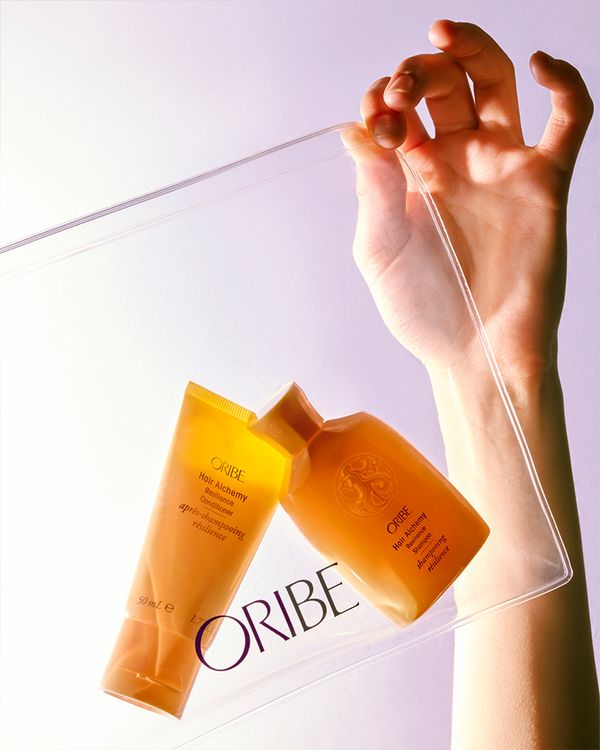 A hand holding a clear pouch with Oribe's Hair Alchemy shampoo and conditioner bottles